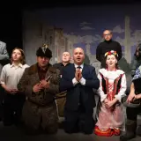 Aktorzy na scenie, w strojach z epoki, tworzą teatr ciut frapujący. W tle malowana miejska panorama.