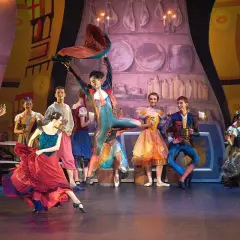 Tancerka w czerwonej sukni wiruje na scenie. W tle grupa postaci w barwnych strojach, jakby z bajki, gdzie teatr nie ma sobie równych.