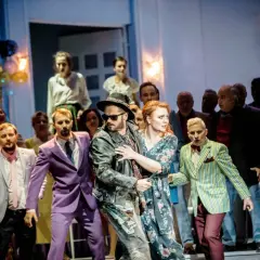 Na scenie opery Manru, bohaterka z rudymi włosami w kwiecistej sukni obejmuje mężczyznę w skórzanej kurtce.