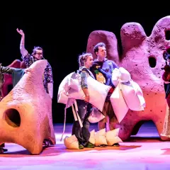 Aktorzy w barwnych strojach, niosący poduszki, na tle scenografii przypominającej gigantyczne, różowe formacje. To fragment spektaklu z repertuaru teatru lalki.