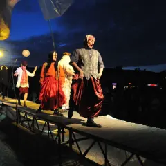Aktorzy w barwnych strojach na scenie. W tle nocne niebo i widzowie. Spektakl "Teatr ósmego dnia" trwa.