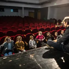 Dzieci słuchają opowieści na scenie, siedząc przed rzędem czerwonych foteli. To część repertuaru teatru Zagłębia.