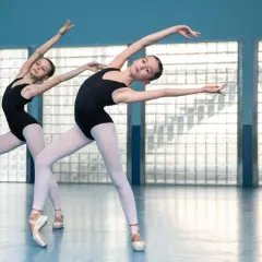 Dwie młode tancerki baletowe w czarnych trykotach i białych rajstopach, stojące na czubkach palców, wykonują elegancką choreografię.