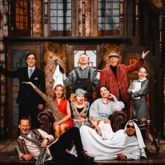 Aktorzy w teatralnej scenografii, jakby w teatrze bez rzędów, pozują do zdjęcia. Wszyscy uśmiechnięci, w barwnych strojach, tworzą radosną atmosferę.