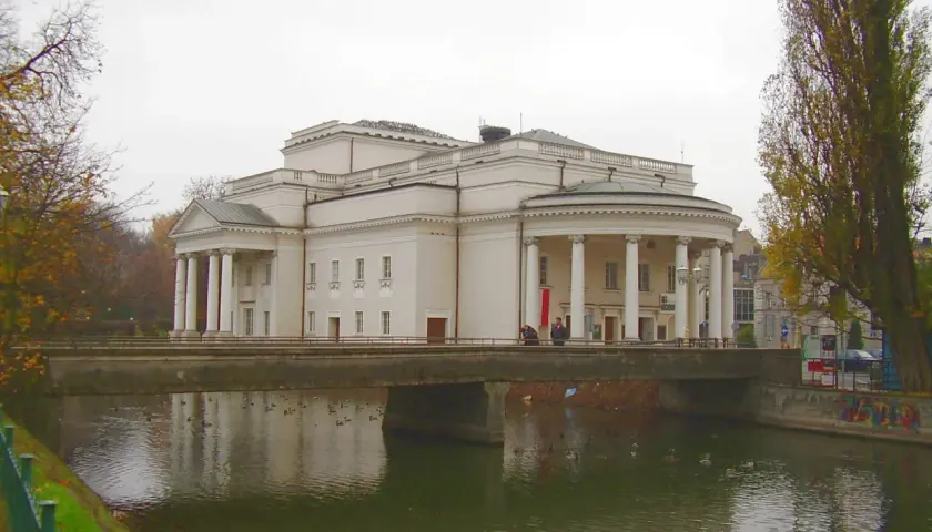 Teatr Kalisz zaprasza na spektakle. Imponujący budynek z kolumnadą nad rzeką, jesienne drzewa.