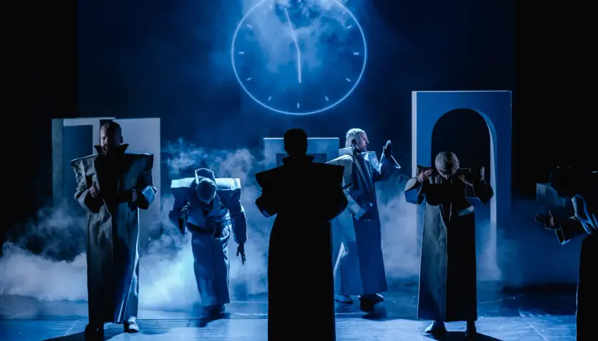 Postacie w futurystycznych strojach na scenie Teatru Arlekin w Łodzi. W tle świecący zegar.