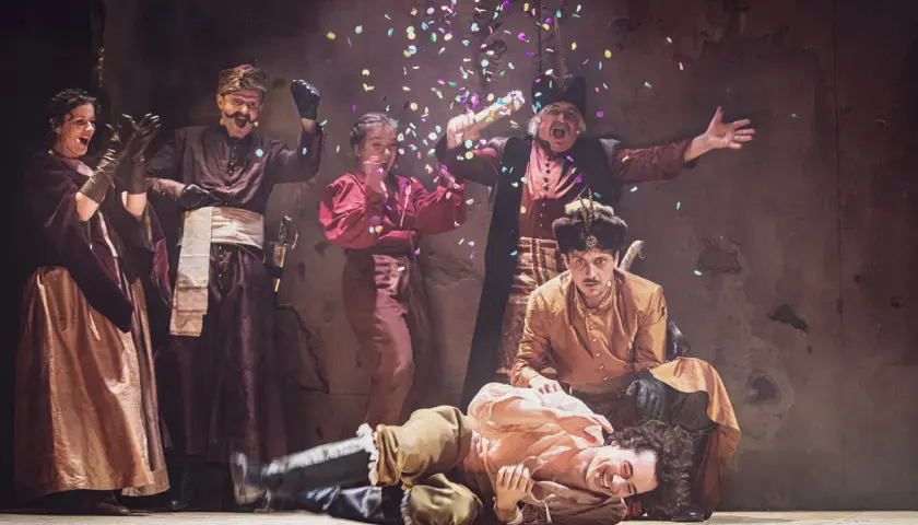 Aktorzy w strojach ludowych świętują na scenie, sypiąc konfetti. To fragment repertuaru teatru ludowego.