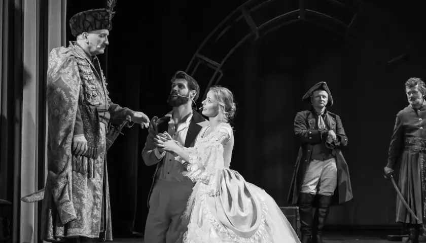 Aktorzy w historycznych strojach na scenie. W centrum młoda para, obok nich starszy mężczyzna w bogatym stroju. To piękny **teatr u przyjaciół**.