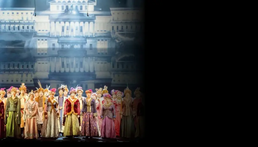 Chór w barwnych strojach historycznych na tle pałacowej scenografii. Sprawdź opera warszawa repertuar!