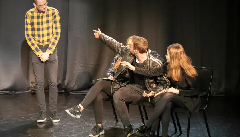 Młodzi aktorzy ćwiczą scenę. Jeden z nich wskazuje coś na widowni, jakby to był teatr na bruku.