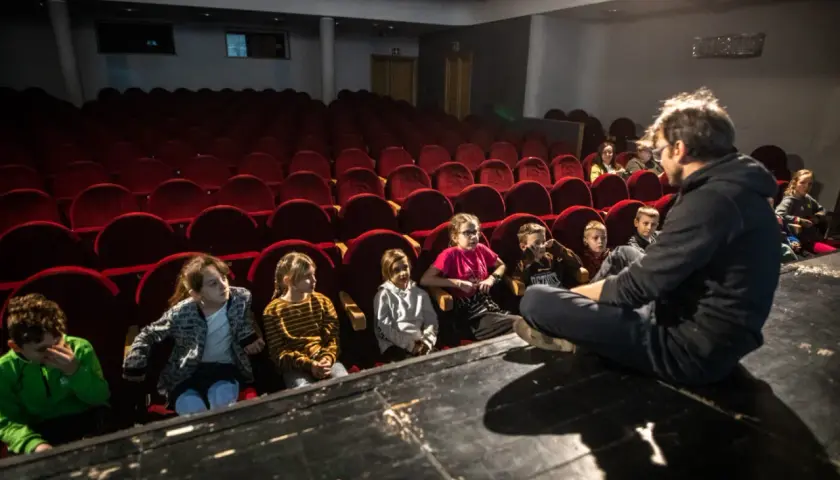 Dzieci słuchają opowieści na scenie, siedząc przed rzędem czerwonych foteli. To część repertuaru teatru Zagłębia.