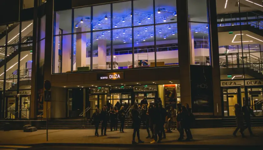 Teatr Muzyczny im. Danuty Baduszkowej nocą. Grupa ludzi czeka przed wejściem, zapowiadając nadchodzące wydarzenia.