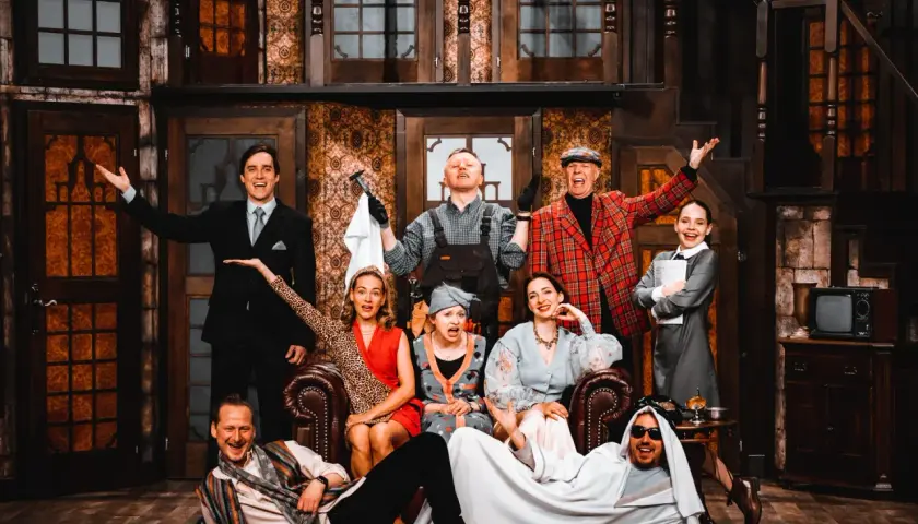 Aktorzy w teatralnej scenografii, jakby w teatrze bez rzędów, pozują do zdjęcia. Wszyscy uśmiechnięci, w barwnych strojach, tworzą radosną atmosferę.