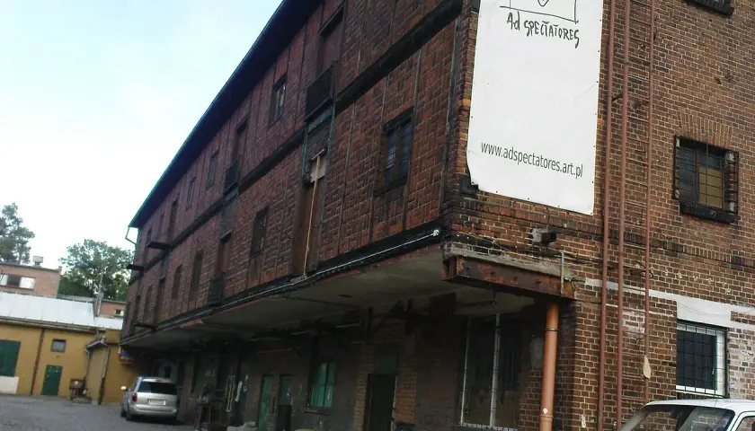 Stara ceglana kamienica z banerem "Ad Spectatores". Obok samochody i żółte krzesła.