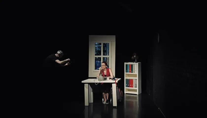 Scena w **teatrze Kana Szczecin**: kobieta przy biurku rozmawia przez telefon, mężczyzna w tle filmuje.