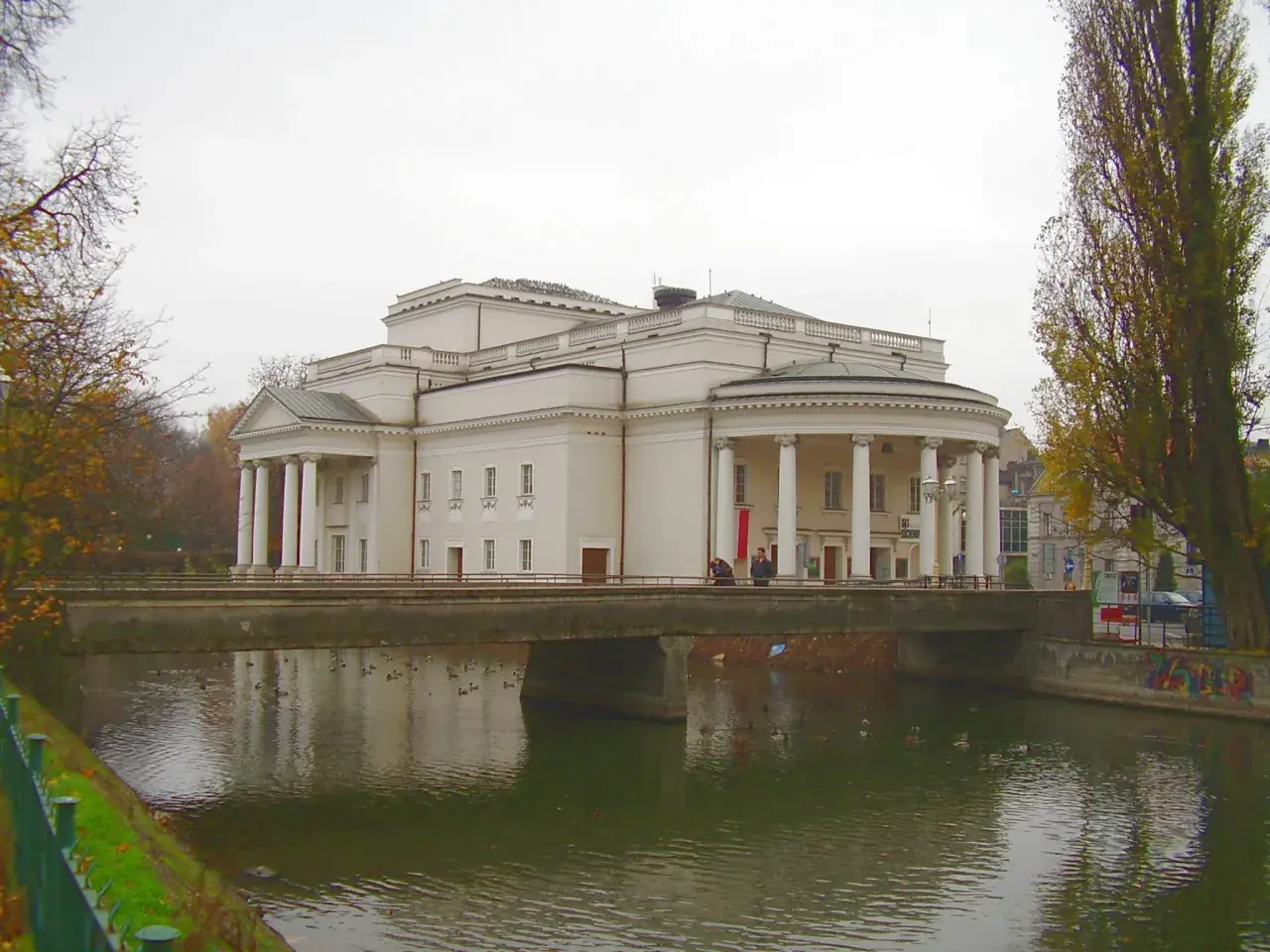 Teatr Kalisz zaprasza na spektakle. Imponujący budynek z kolumnadą nad rzeką, jesienne drzewa.
