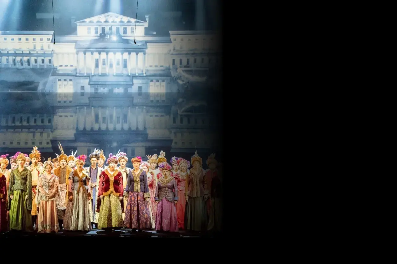 Chór w barwnych strojach historycznych na tle pałacowej scenografii. Sprawdź opera warszawa repertuar!
