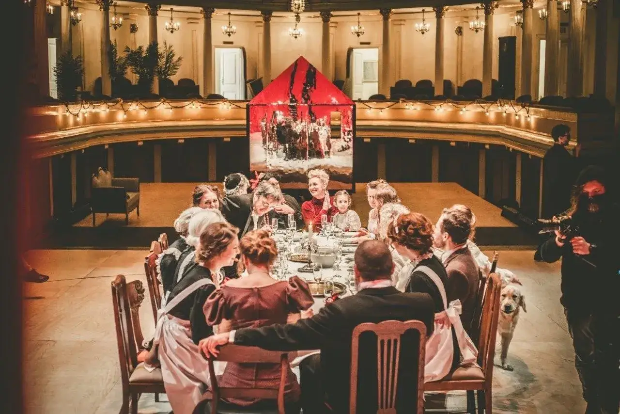 Scena z bankietem i czerwonym domkiem w tle. Sprawdź repertuar Teatru Modrzejewskiej w Legnicy, by zobaczyć więcej takich widowisk.