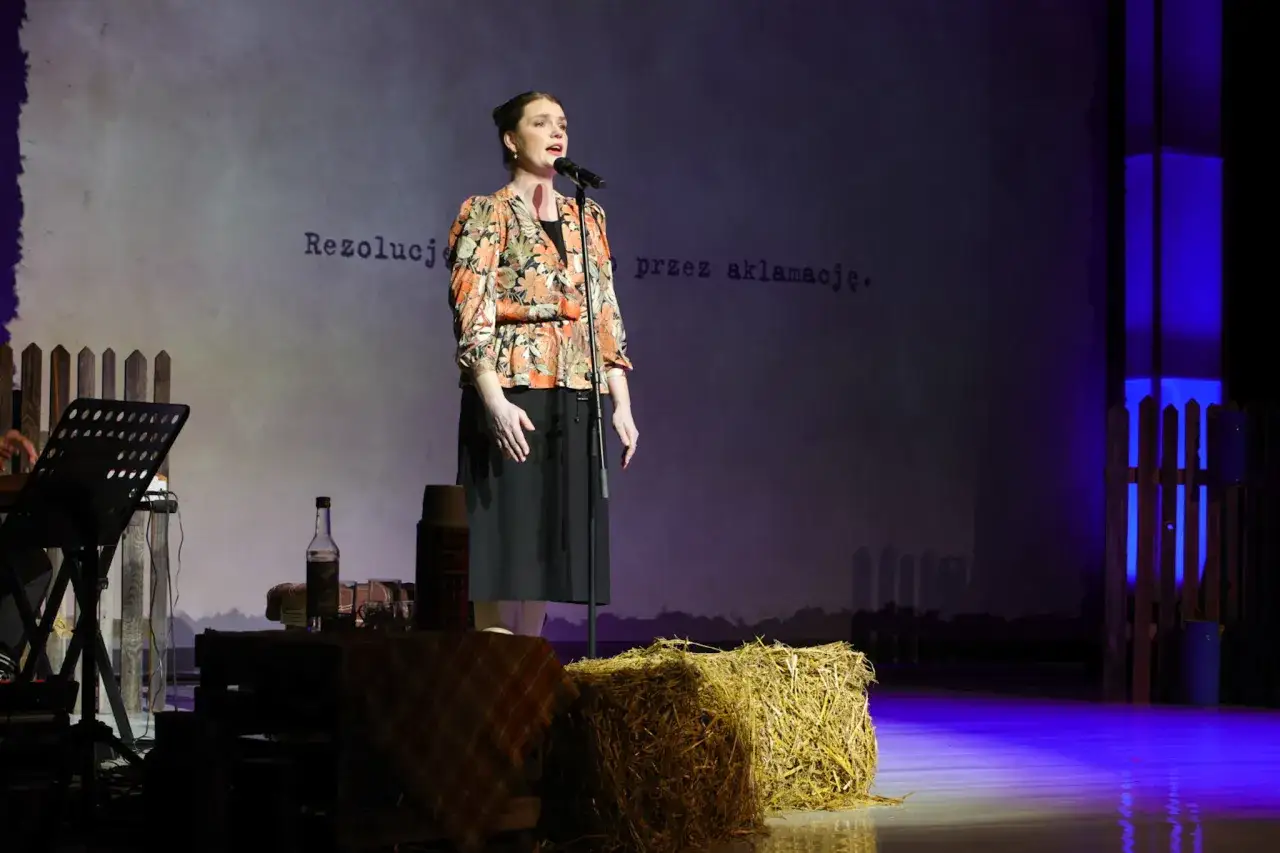 Kobieta śpiewa na scenie, przypominając o polskim teatrze w podziemiu. Na scenie słoma, butelka i termos.