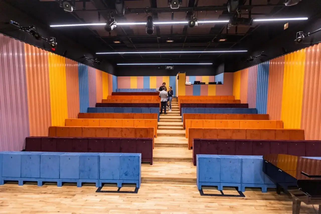 Kolorowe siedziska i ściany tworzą nowoczesną przestrzeń, idealną dla teatru ludowego w Krakowie. Trzy osoby rozmawiają na schodach.